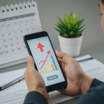 Pessoa observando gráfico de score de crédito positivo em smartphone, com calendário de três meses ao fundo e documentos financeiros.